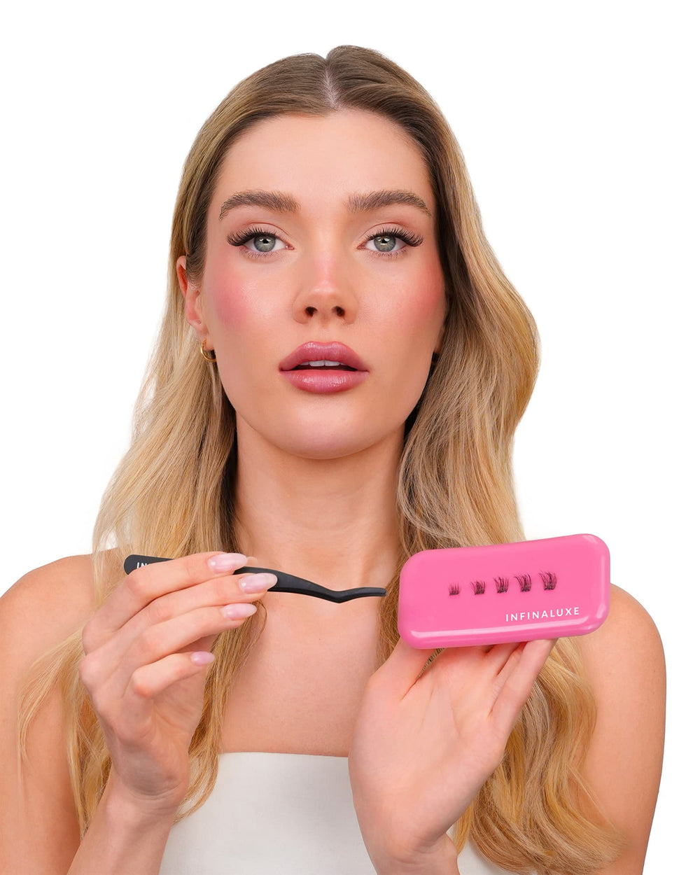 A blonde woman holding a pink Infinaluxe lash extension kit, including a small pink case and a black spatula, against a solid white background.