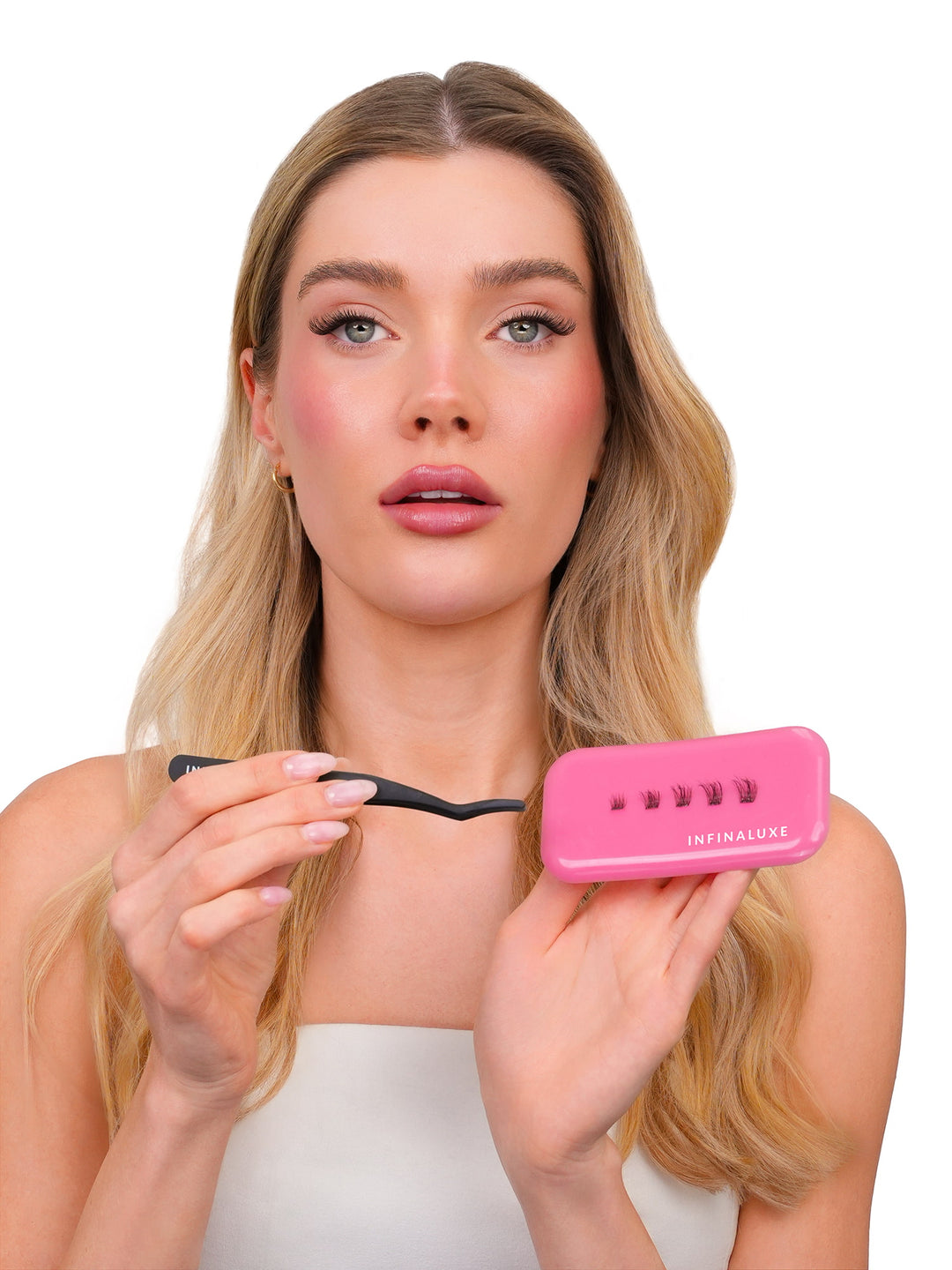 A blonde woman holding a pink Infinaluxe lash extension kit, including a small pink case and a black spatula, against a solid white background.